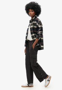 Geruite overhemdbloes in marineblauw, wit en bordeaux; witte t-shirt; zwarte cargobroek met ritsdetails; witte sneakers met zwarte strepen.