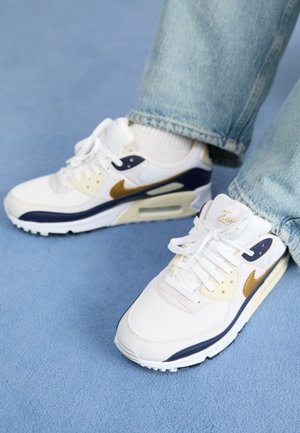 Baskets blanches avec des accents bleu marine et dorés, en matériaux en mesh et cuir, côtés perforés, lacets plats et une unité de coussin d'air visible.
