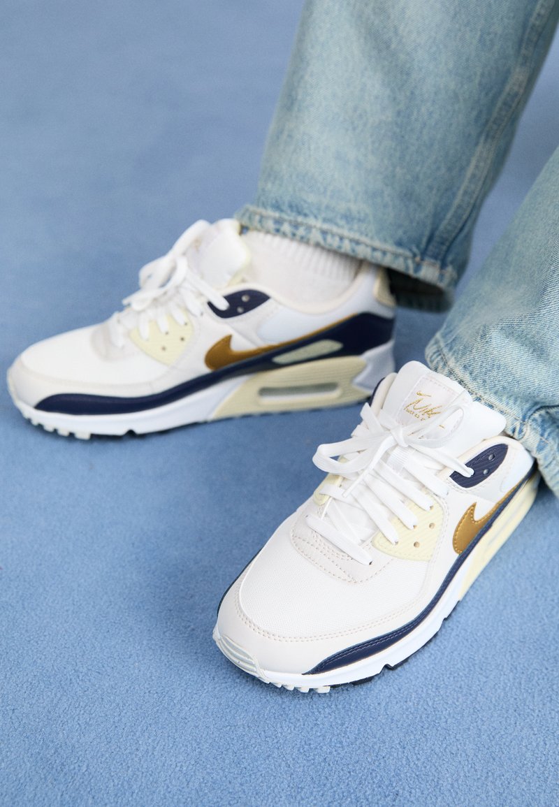Zapatilla blanca con acentos en marino y dorado, materiales de malla y cuero, lados perforados, cordones planos y una unidad de amortiguación de aire visible.