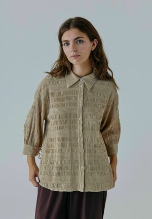 Beige, struktureret skjorte med lodrette plisseer, knaplukning foran og spredt krave. Har trekvart puffærmer og en løs pasform.