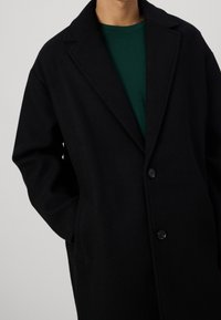Schwarzer Wollmantel mit Reverskragen, zwei Knöpfen und Seitentaschen, getragen über einem grünen Hemd. Die Textur wirkt glatt.