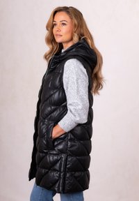 Woman with long blonde hair wearing a black quilted sleeveless jacket and gray sweater, standing in profile with hands in pockets.