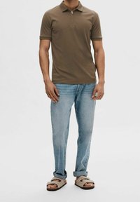 Olivgrön polo t-shirt med halv dragkedja, i kombination med ljusblå jeans och bruna sandaler. T-shirten har korta ärmar och en klassisk krage.