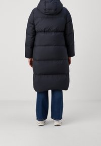 Person, der einen langen schwarzen Daunenmantel mit Kapuze, blaue Jeans und weiße Sneaker trägt, steht mit dem Rücken zur Kamera vor einem schlichten Hintergrund.
