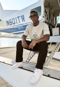 Camiseta beige claro con el texto "inocente hasta que se demuestre lo contrario", combinada con pantalones negros y zapatillas blancas, sentado sobre un ala de avión.
