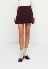 Vrouw in een hoog taille, geplisseerde bordeauxrode corduroy rok, een witte blouse met lange mouwen, beige sokken en bordeauxrode ballerina's, staande op een witte vloer.