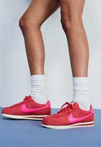 Piros és rózsaszín Nike sportcipők sima felsőrésszel, kiemelkedő oldalsó logóval és mintás gumitalppal rendelkeznek. Fehér zoknival viselve.