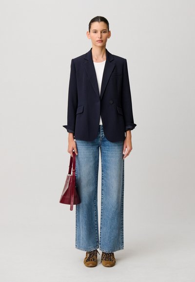 Mujer de pie con blazer azul marino, camisa blanca, vaqueros anchos azules, zapatillas marrones y sosteniendo un bolso rojo oscuro sobre un fondo liso.