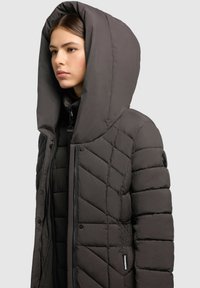 Joven mujer que lleva un abrigo acolchado gris oscuro con capucha grande, abrochado y cerrado, de perfil hacia la izquierda sobre un fondo claro y liso.