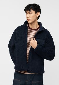 Νέος άνδρας που φοράει μπλε σκούρο fleece μπουφάν πάνω από ριγέ πουκάμισο σε βουργουνδί και μπεζ, με σκουρόχρωμο τζιν παντελόνι, κοιτάζει προς τα δεξιά.