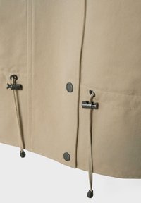 Ourlet de veste en tissu beige avec cordons ajustables comportant des boutonnières métalliques et des boutons-pression le long de la fermeture frontale.