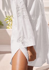 Chemise en lin blanche avec broderies florales, manches longues, détails de boutons et fente sur le côté. Texture douce, design léger et coupe décontractée.
