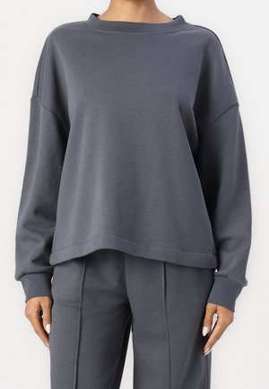 Persona con sudadera gris oscuro de corte holgado y pantalones a juego, de pie con las manos relajadas a los lados, sobre un fondo blanco.