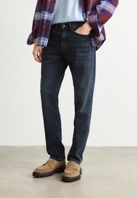 Persona con jeans azul oscuro, mocasines color beige, camisa azul clara y chaqueta a cuadros roja y azul, de pie sobre un suelo claro.