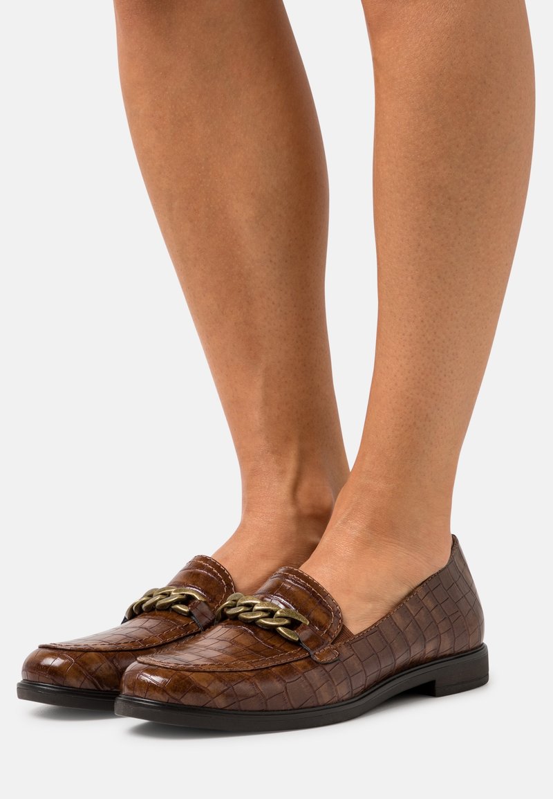 Mocassins en cuir marron à motif crocodile avec un détail de chaîne dorée portés pieds nus avec un ton de peau brun clair.