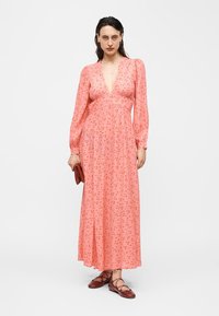 Femeie purtând o rochie maxi cu mâneci lungi, cu imprimeu floral roz și un decolteu adânc în V, ținând o poșetă roșie mică și purtând pantofi roșii cu șireturi.