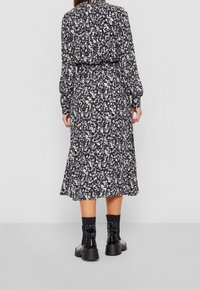 Frau in einem langärmeligen Midikleid mit einem schwarz-weißen und grauen Blumenmuster, kombiniert mit schwarzen Stiefeln bis zur Mitte der Wade, von hinten gezeigt.