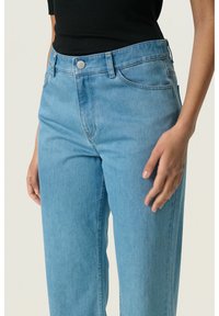 Jeans de denim azul claro con ajuste recto, presentan un cierre de botón metálico, pasadores para el cinturón y bolsillos frontales. Textura suave y costuras mínimas.