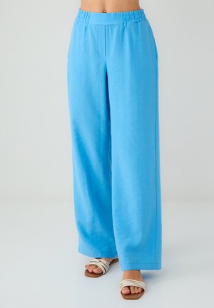 Donna che indossa pantaloni ampi di un brillante blu e sandali piatti bianchi con cinghie, in piedi su un pavimento bianco con uno sfondo semplice.