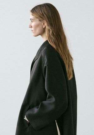 Sort oversized frakke med glat tekstur, bred krave og sidelommer. Model med langt, glat hår, sat op mod en neutral baggrund.