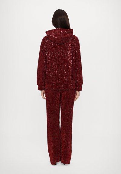 Red sequined tracksuit featuring a hooded jacket and wide-leg pants. The fabric is shiny with a textured surface due to sequins.