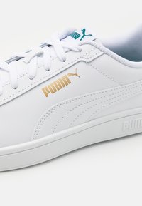 Sneaker in pelle bianca con una texture liscia, caratterizzate da un branding oro e teal, lacci rotondi e colletto imbottito.