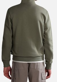 Persona che indossa una felpa a maniche lunghe di colore verde oliva e pantaloni cargo beige chiaro, in piedi rivolto di spalle su uno sfondo bianco uniforme.