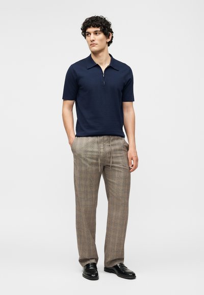 Άνδρας με σγουρά μαλλιά που φοράει μπλε κοντομάνικο πουκάμισο με φερμουάρ, μπεζ καρό παντελόνια και μαύρα δερμάτινα παπούτσια, στέκεται με τα χέρια στις τσέπες.