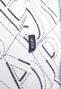Tessuto bianco con cuciture astratte blu navy, caratterizzato da un piccolo etichetta nera con la scritta "JOOP!" sul bordo. Texture liscia e design moderno.