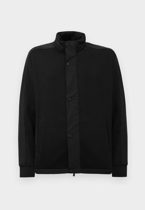 Svart jakke med høy krage, trykknapper og kontrasterende stoffpaneler. Har to sidelommer og ribbestrikkede ermer for en tettsittende look.