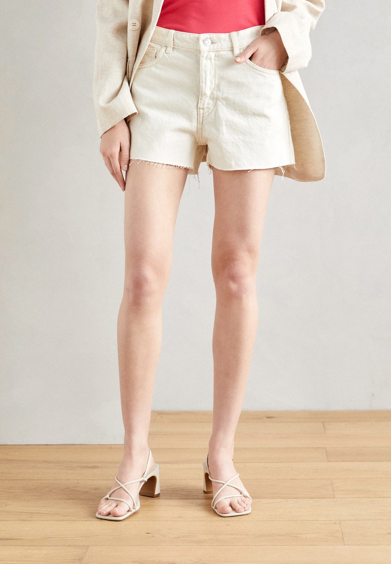 White denim shorts with frayed hem, paired with a red top and beige strappy heels. Smooth texture, fitted design, and casual look.