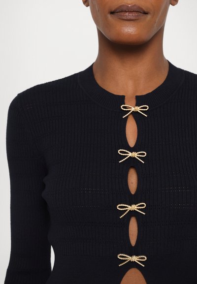 Crna pletiva majica s rebrastom teksturom, okruglim izrezom i zlatnim mašnicama na ovalnim izrezima duž prednje strane.