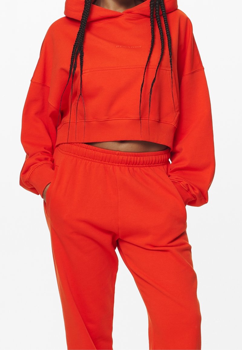 Personne portant un sweat à capuche court orange vif et un pantalon de survêtement assorti, les mains dans les poches, cheveux tressés tombant devant.