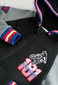 Schwarze und graue Kapuzenjacke mit Reißverschluss, die ein gesticktes Motorrad-Motiv, den Text "181" und gerippte Bündchen mit blauen, roten und weißen Streifen aufweist.