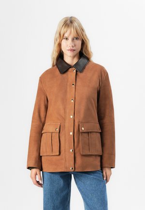 Donna che indossa una giacca scamosciata marrone con colletto marrone scuro, bottoni a pressione dorati, tasche anteriori con patta e jeans blu su sfondo bianco.