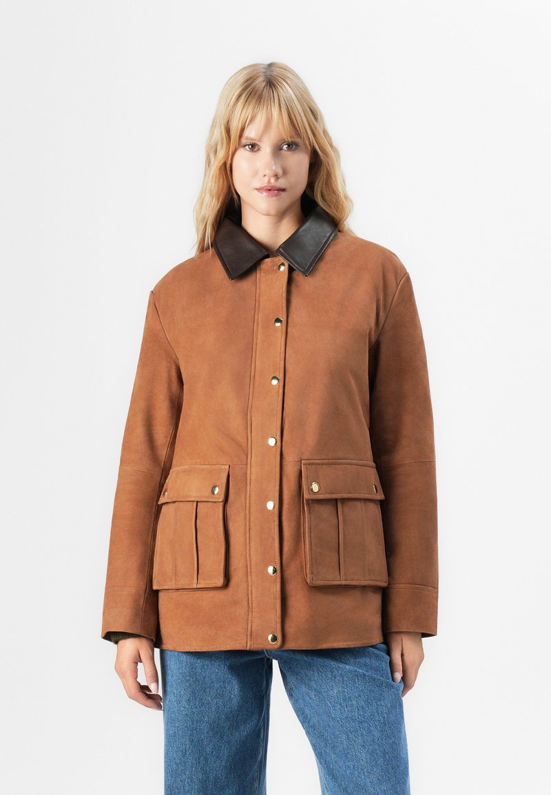 Donna che indossa una giacca scamosciata marrone con colletto marrone scuro, bottoni a pressione dorati, tasche anteriori con patta e jeans blu su sfondo bianco.