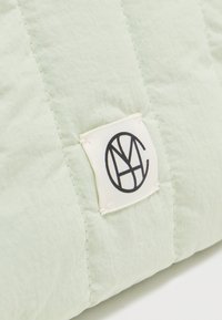 Υφασμα σε ανοιχτό πράσινο με κάθετες ραφές και μια λευκή ορθογώνια ετικέτα που φέρει μαύρο κυκλικό γεωμετρικό λογότυπο.