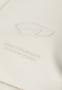 White fabric with a textured surface. Features a raised logo and embossed coordinates reading "55°40'34"N 12°34'06"E."