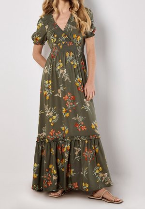 Femme portant une robe longue vert olive avec un imprimé floral orange, jaune et crème, manches bouffantes et ourlet à volants, associée à des sandales beiges.