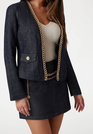 Vrouw die een donker denim jack en rok draagt met gouden kettingaccent, gecombineerd met een wit geribd mouwloos topje.