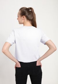 Bílá krátká tričko s krátkým rukávem zobrazené zezadu, má kulatý výstřih a volný střih. Model má na sobě černé kalhoty.