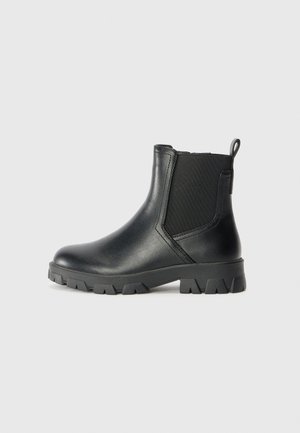Bottine noire en cuir avec panneaux latéraux élastiques, languette à l'arrière et semelle épaisse à crampons sur fond clair.