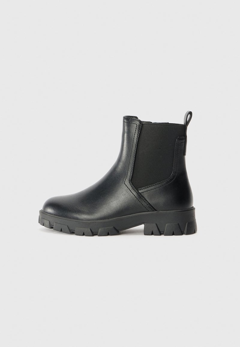 Bottine noire en cuir avec panneaux latéraux élastiques, languette à l'arrière et semelle épaisse à crampons sur fond clair.