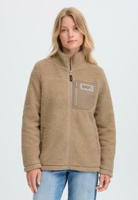 Femme portant une veste en polaire beige avec fermeture éclair à l'avant et poche poitrine, associée à un jean bleu clair, se tenant devant un fond uni.
