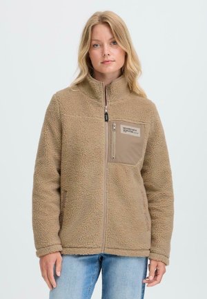Vrouw in een beige fleecejack met een rits aan de voorkant en borstzak, gecombineerd met lichtblauwe jeans, staande tegen een effen achtergrond.