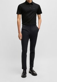 Homme portant un polo noir à manches courtes, un pantalon slim noir et des mocassins en cuir noirs sur un fond clair.