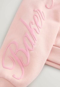 Różowy materiał bluzy z uniesionym napisem "Baker" w dopasowanym odcieniu różu. Tekstura jest gładka, a końce rękawów mają ryflowane mankiety.