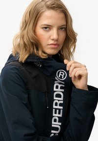 Granatowa kurtka z czarnymi akcentami, z wyraźnym białym logo w okolicy zamka błyskawicznego oraz kapturem z regulowanymi trokami.