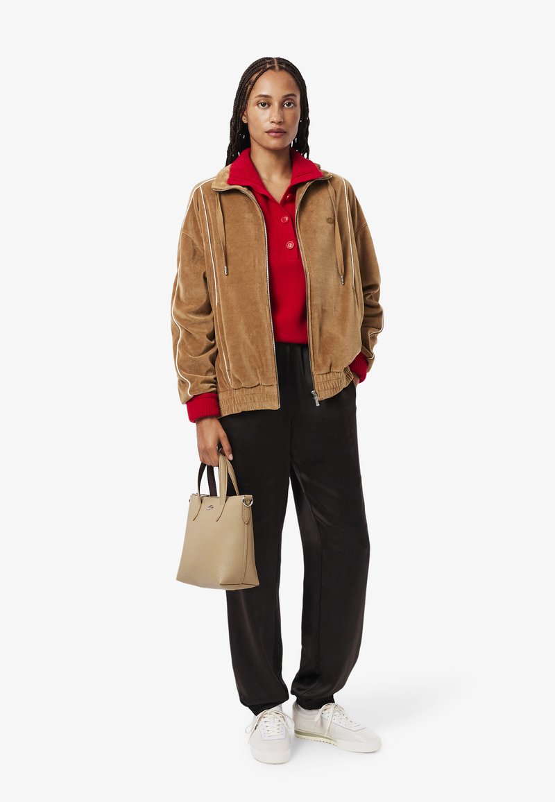 Giubbotto bomber in suede marrone, polo rossa, pantaloni sportivi neri e sneakers bianche. Tieni una piccola borsa beige. Outfit casual con colori a contrasto.