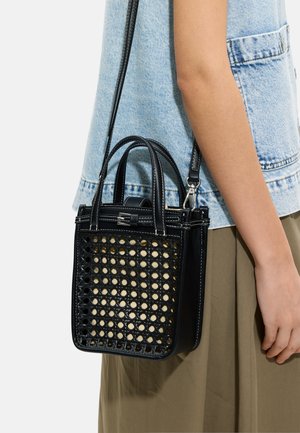 Czarna pleciona skórzana torebka z dwoma uchwytami i odpinanym paskiem na ramię, noszona na ukos przez osobę w kamizelce jeansowej i beżowej spódnicy.